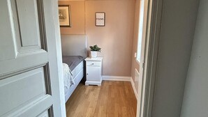 Un couloir étroit mène à une chambre où se trouvent un lit, une table de chevet et un tableau encadré au mur.