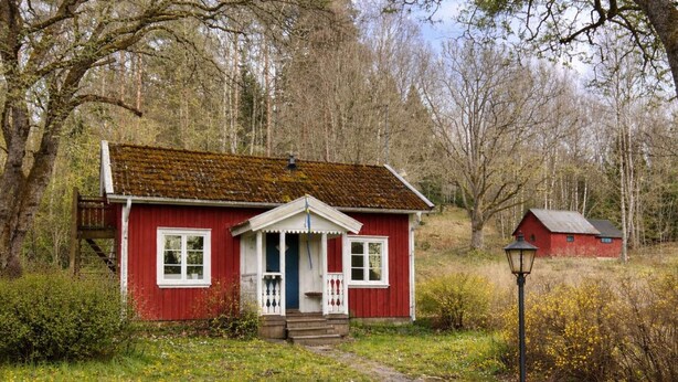 Une maison en bois rouge, dotée d’un porche, d’un petit escalier et d’un lampadaire, située dans un cadre naturel.