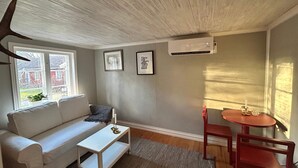 Une pièce avec un canapé blanc, une petite table blanche, une chaise rouge, une autre petite table et une fenêtre donnant sur des maisons.