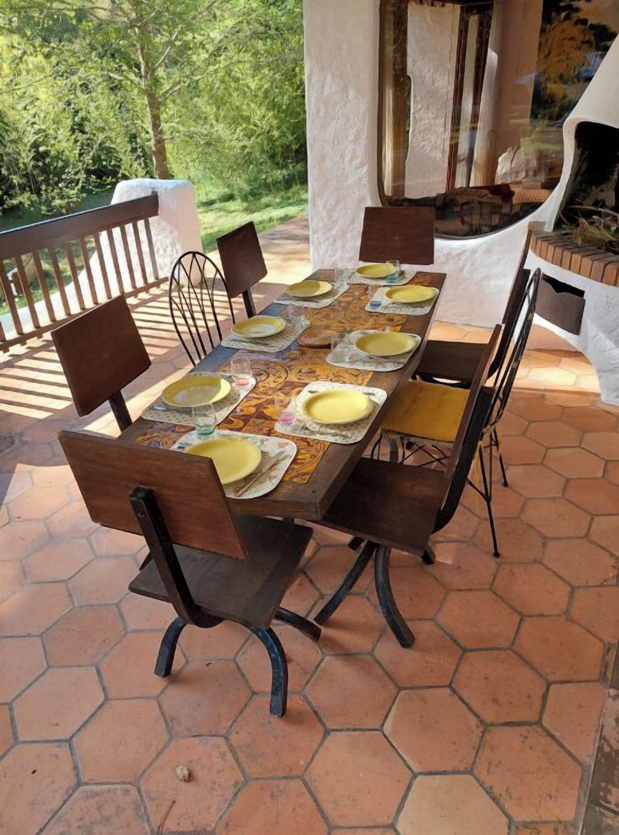 An outdoor dining area with a hexagonal tiled floor, a wooden table set with plates and cutlery, and metal chairs.