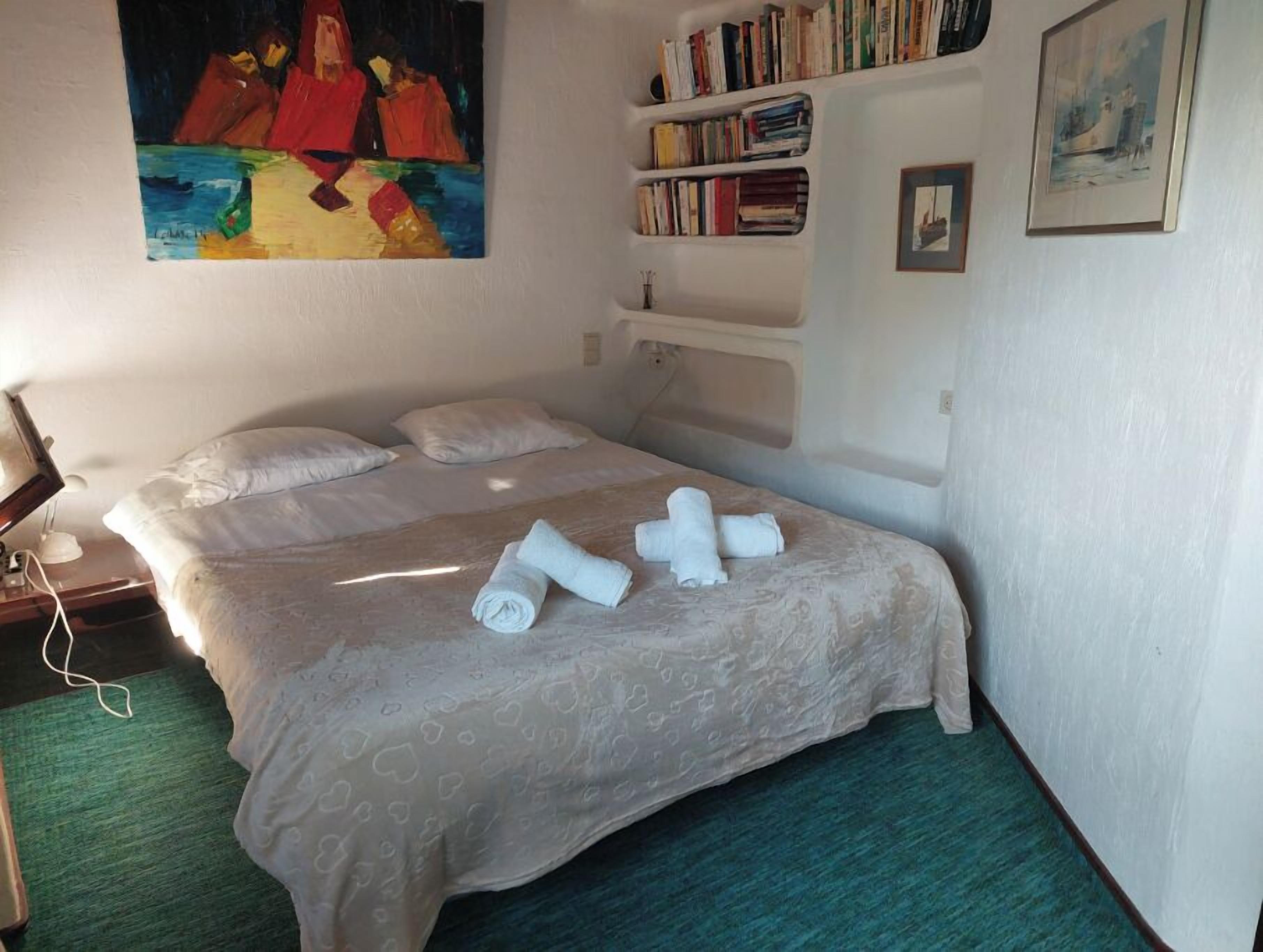 A neatly made bed with rolled towels, a bookshelf with books, and a painting on the wall.