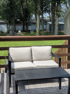 A patio with a cushioned bench and a table, surrounded by a wooden deck and a grassy yard. - The Cozy Retreat
Comfort, Fun & Convenience

 (Windsor Heights)
