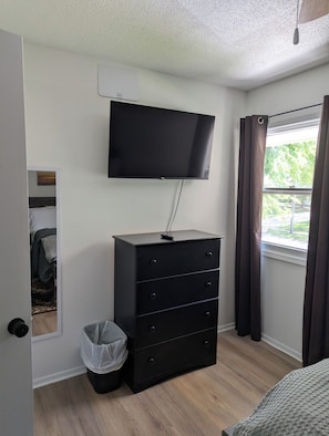 A bedroom with a flat-screen TV mounted on the wall, a black dresser, a trash bin, and a window with curtains. - The Cozy Retreat
Comfort, Fun & Convenience
(Windsor Heights)
