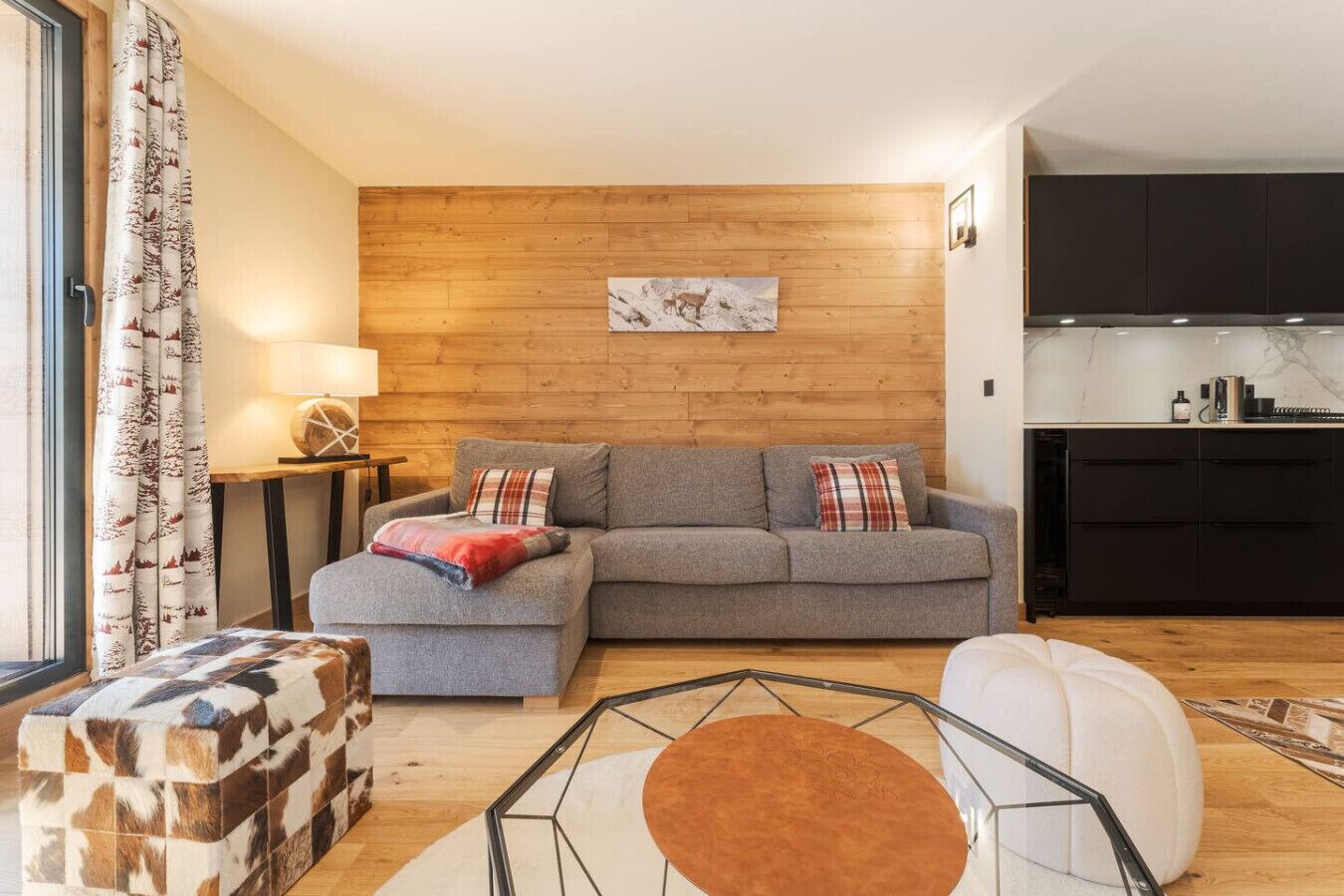 A living room with a grey sofa, a wooden wall, a kitchen area, and a patterned ottoman.