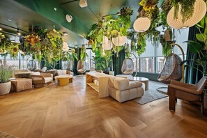 Un amplio salón con suelo de madera, asientos cómodos, plantas colgantes y grandes ventanales con vistas a la ciudad.