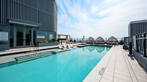 Una piscina en la azotea con sillas de descanso, un edificio moderno con líneas verticales y un cielo despejado.