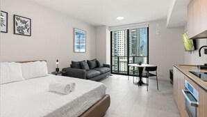 Habitación de hotel moderna con una cama grande, un sofá, una pequeña mesa de comedor y vistas de rascacielos.