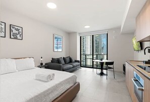 A modern hotel room with a large bed, a sofa, a small dining table, and a view of high-rise buildings. - Downtown Studio 1409 | Rooftop Pool & City Views (Miami)