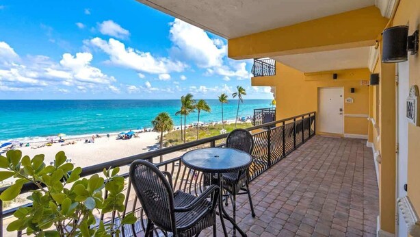 A balcony with a view of the beach and ocean, a round table, and two chairs. - Oceanfront 2BR/2BA · Pool, Free Parking + Views (Hollywood)