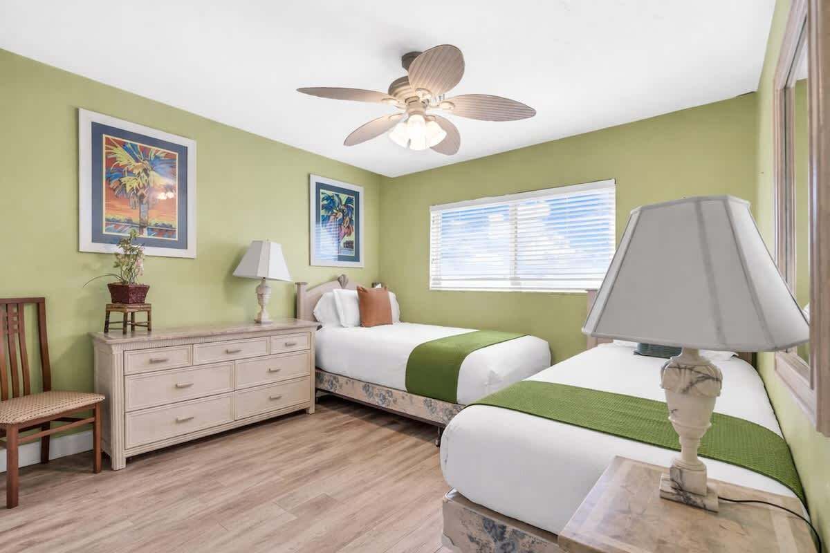 Une chambre avec deux lits, un ventilateur de plafond, une commode, une chaise et des œuvres d’art accrochées aux murs.