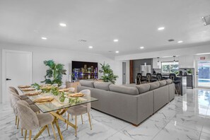 Un'area pranzo moderna con tavolo in vetro, sedie imbottite, un divano grigio e una cucina sullo sfondo.