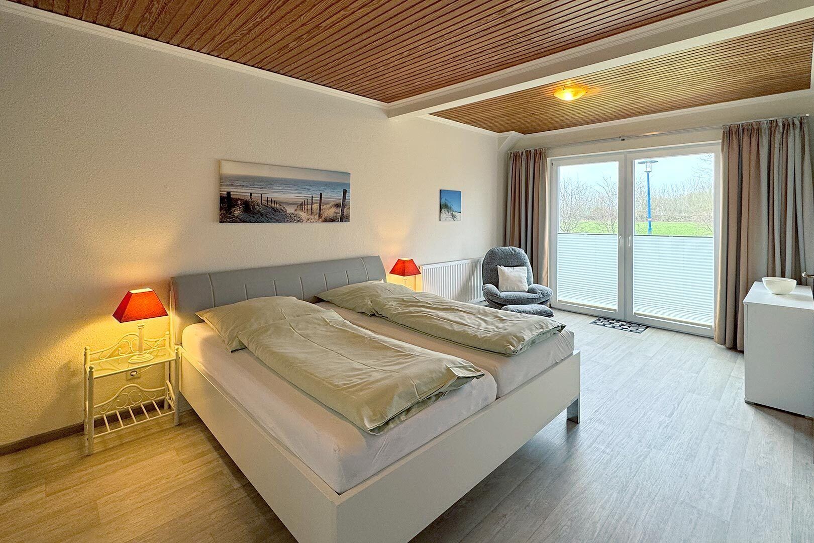 A bedroom with a bed, two bedside tables with lamps, a chair, a window with curtains, and a painting on the wall.
