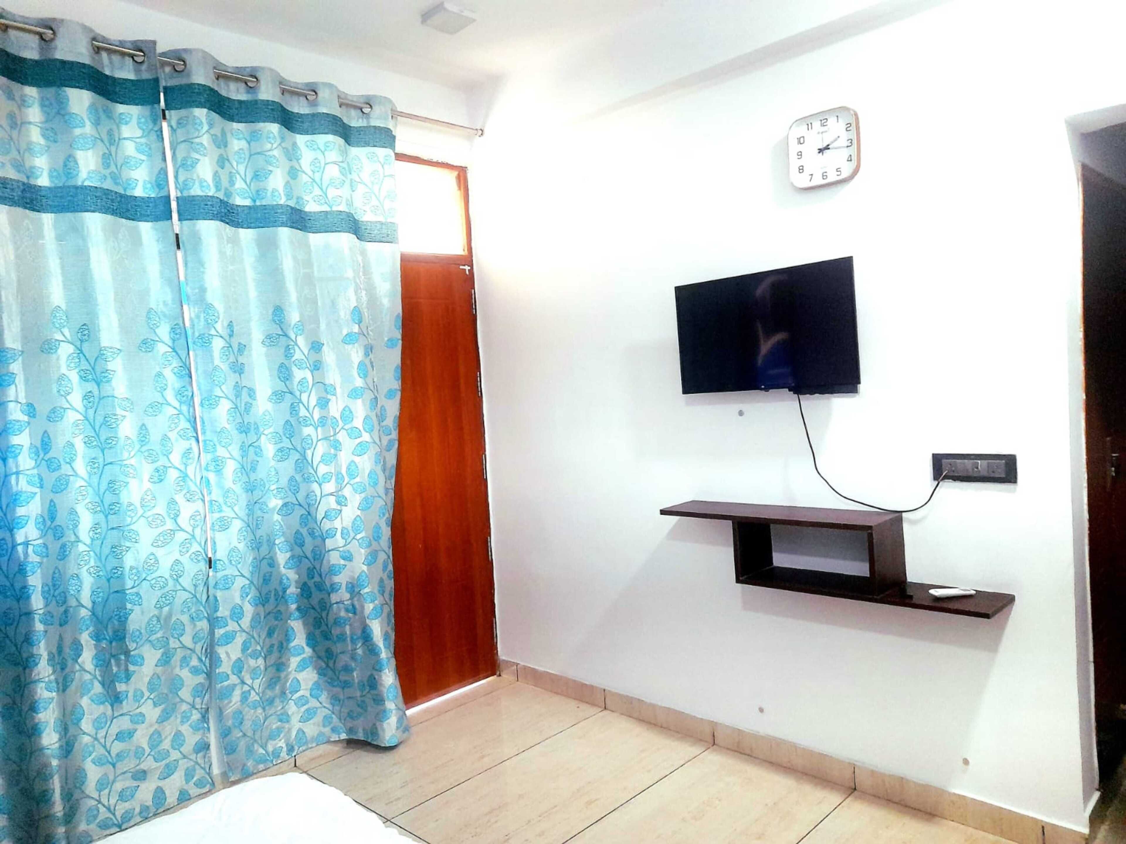 A room with a TV mounted on the wall, a clock, a wooden door, and a curtain with a leaf pattern.