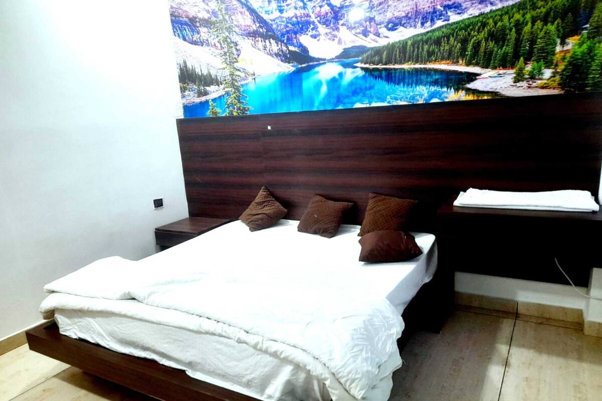 A bedroom with a bed, pillows, a nightstand, a wooden headboard, and a large painting of a mountain landscape.