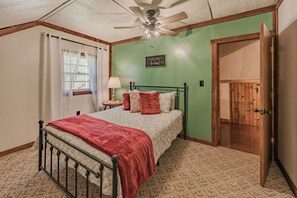 Une chambre à coucher comprenant un lit, un ventilateur de plafond, une fenêtre avec des rideaux, une table de chevet avec une lampe et une porte.