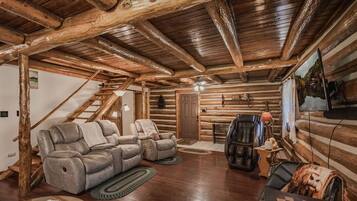 A log cabin interior with a wooden ceiling, a reclining sofa, a leather armchair, a television, and a fireplace.