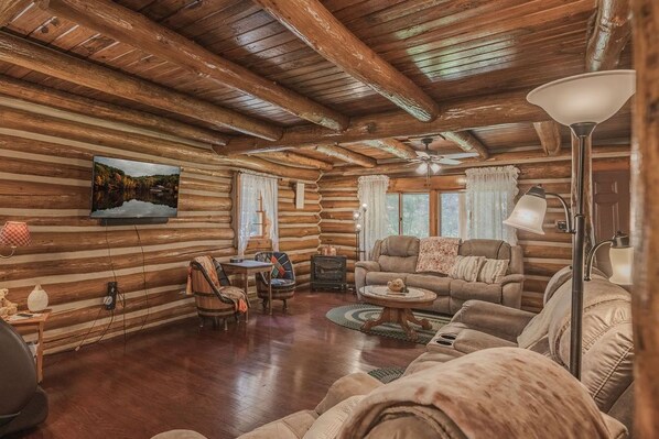 L’intérieur d’une cabane en rondins, avec un plafond en bois, un téléviseur fixé au mur, une cheminée et un coin salon confortable.