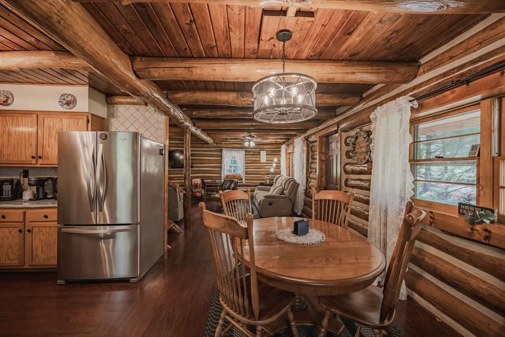 A log cabin interior with a wooden dining table, chairs, a refrigerator, and a hanging light fixture.