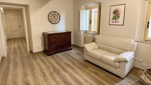 A room with a wooden floor, a white sofa, a wooden dresser, a wall clock, and a window with a view. - CASA AMBRA (Cortona)