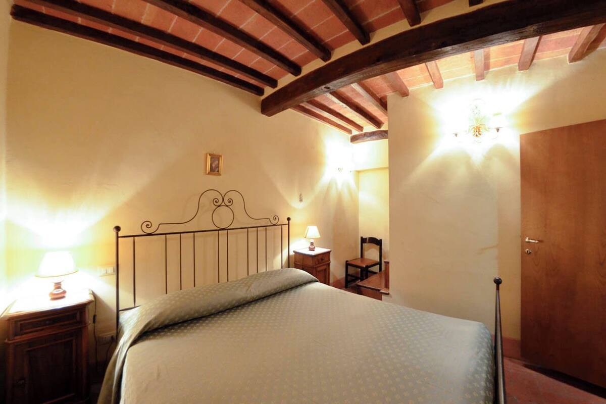 A bedroom with a bed, a nightstand, a chair, a wooden door, and a ceiling with exposed beams.