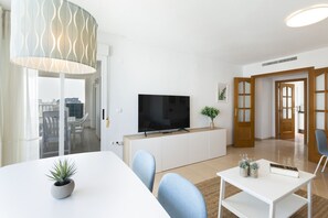 A modern living room with a flat-screen TV, a dining table with chairs, a potted plant, and a large window. - Beautiful flat with mediterranean style and max confornt - only families (Playa de Gandía)