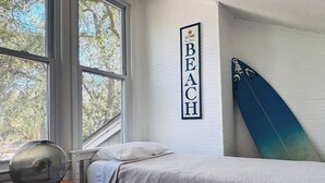 Een slaapkamer met een bed, een surfplank, een bord aan de muur en een raam met uitzicht op bomen.