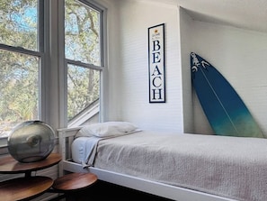 Een slaapkamer met een bed, een surfplank, een bord aan de muur en een raam met uitzicht op bomen.