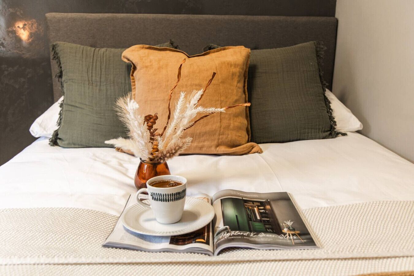 A bed with white bedding, a magazine, a cup of coffee, and decorative pampas grass.