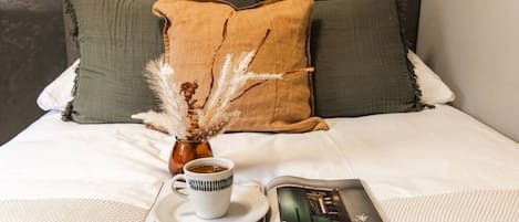 En säng med vita sängkläder, ett tidningsnummer, en kopp kaffe och dekorativt prydnadsgräs.