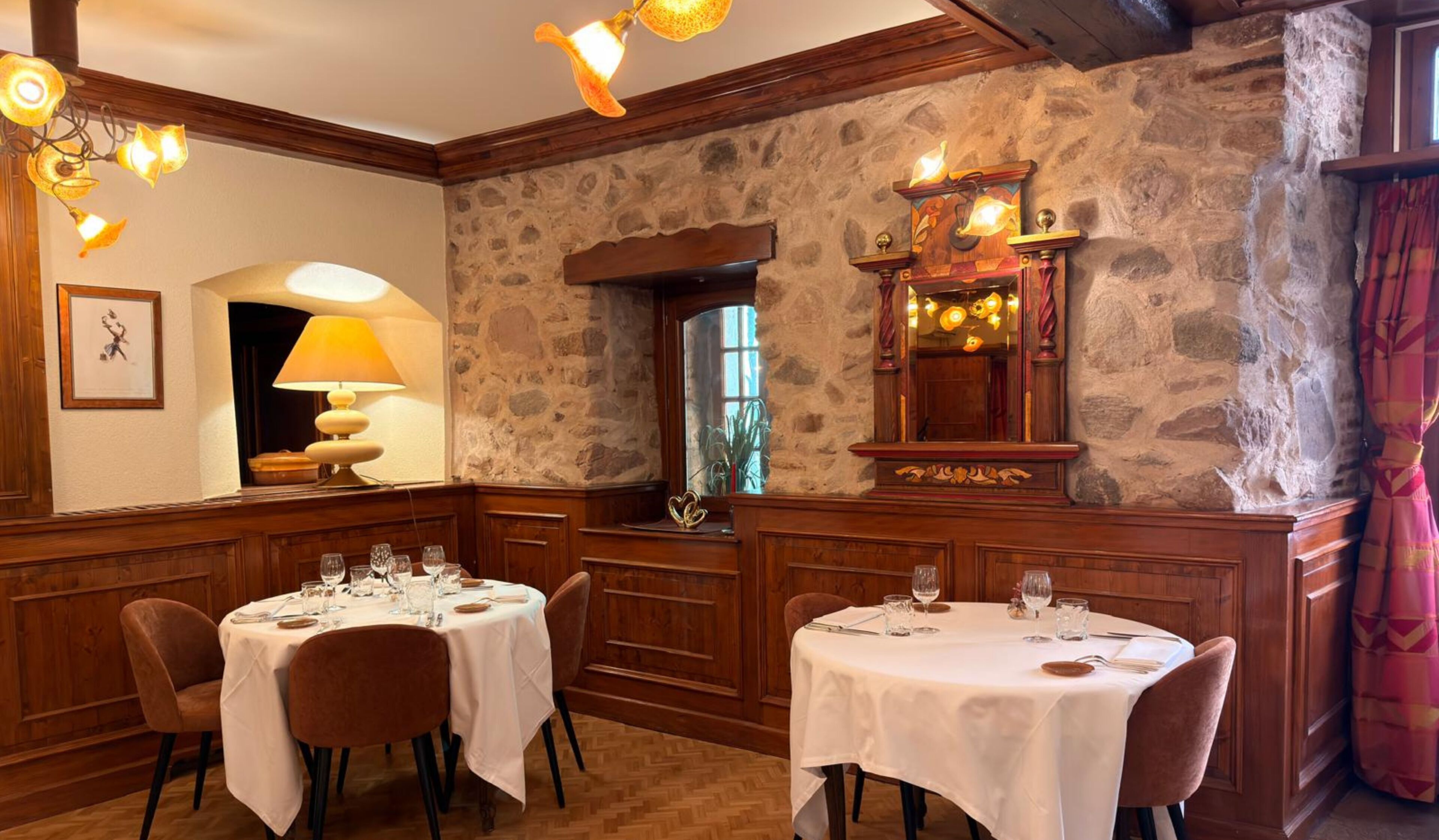 Un comedor con paredes de piedra, paneles de madera y candelabros. Las mesas están dispuestas con manteles blancos y copas de vino.