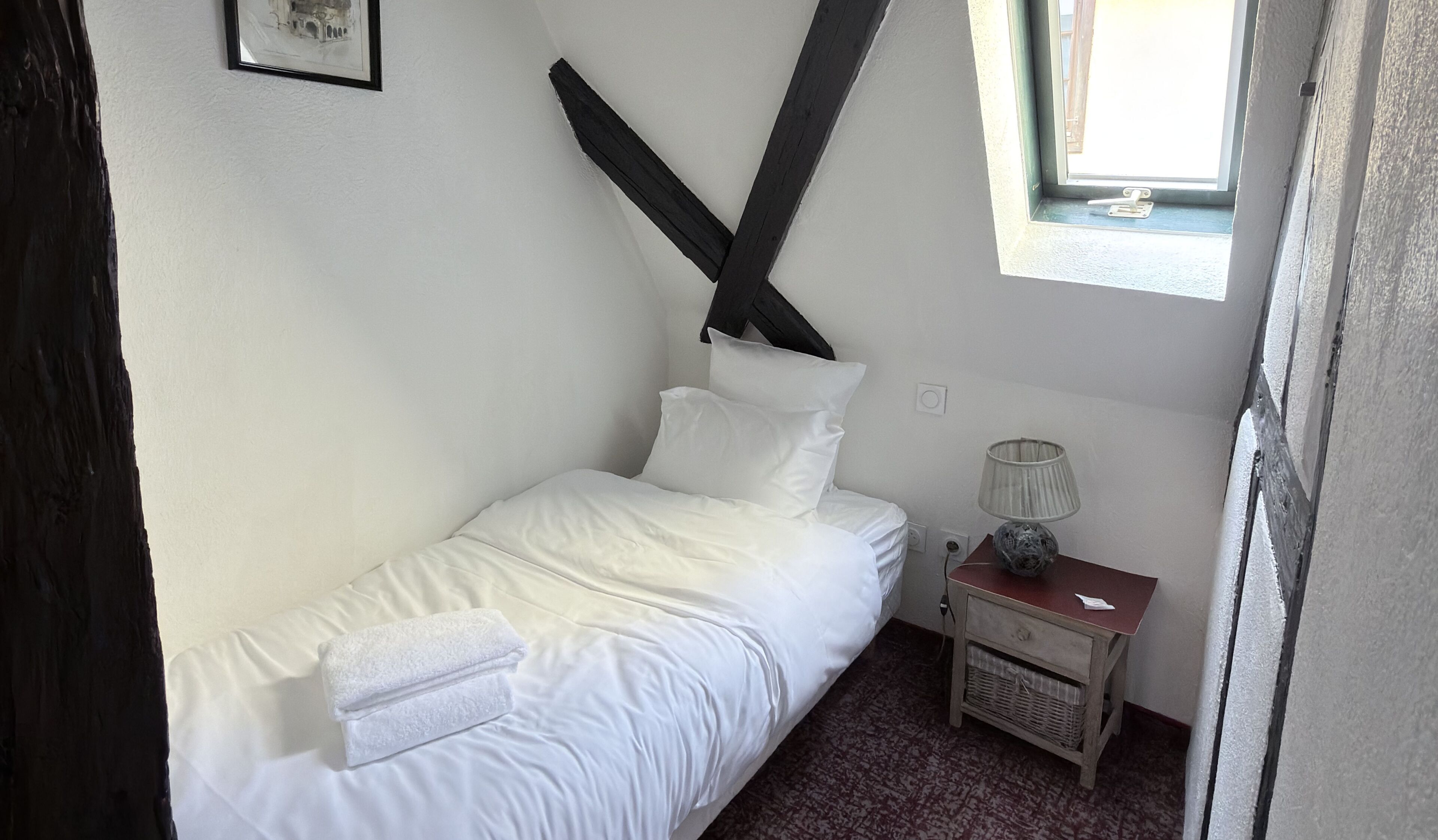 Una cama pequeña y bien hecha con sábanas y almohadas blancas, una lámpara de noche, una mesita de noche de madera y un cuadro en la pared.