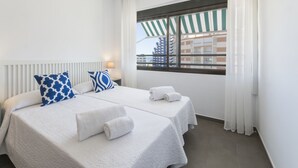 Une chambre d’hôtel avec deux lits, du linge de lit blanc, une fenêtre donnant sur des immeubles et une table de chevet avec une lampe.