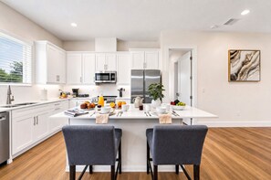 A modern kitchen with white cabinets, stainless steel appliances, a central island with seating, and a dining area with two chairs. - Private Patio | Ground Floor | Near Busch Gardens (Tampa)