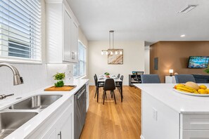 A modern kitchen with white cabinetry, a dining area with a wooden table, and a living space with a TV and seating. - Bright & Modern 3BR Home – Prime Tampa Location (Tampa)