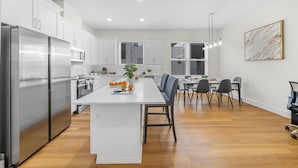 Una cocina moderna con armarios blancos, un refrigerador de acero inoxidable, una isla central con frutas y un comedor con sillas y una mesa.