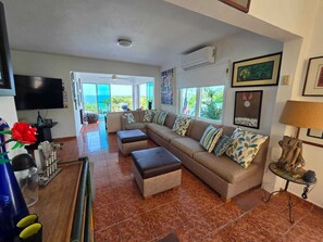 A living room with a large sectional sofa, a television, and a view to the outside. - Bluecove Hideaway Relaxing Retreat - Guarida Azul 3BR  Private Pool + Ocean View (Quebradillas)