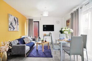 Ein Wohnzimmer mit einer Couch, einem Esstisch mit Stühlen, einem Flachbildfernseher und einem großen Fenster mit Vorhängen.