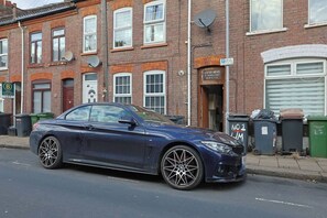 Ein blauer BMW steht auf einer Wohnstraße mit Ziegelhäusern, Satellitenschüsseln und Mülltonnen.