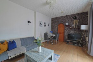 Ein Wohnzimmer mit Sofa, Couchtisch, Esstisch und Stühlen. Eine Steinwand, ein Fernseher und eine Lampe sind ebenfalls vorhanden.
