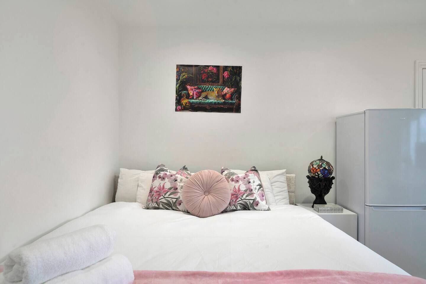 A bedroom with a bed, pillows, a wall art piece, a small table with a decorative item, and a refrigerator.