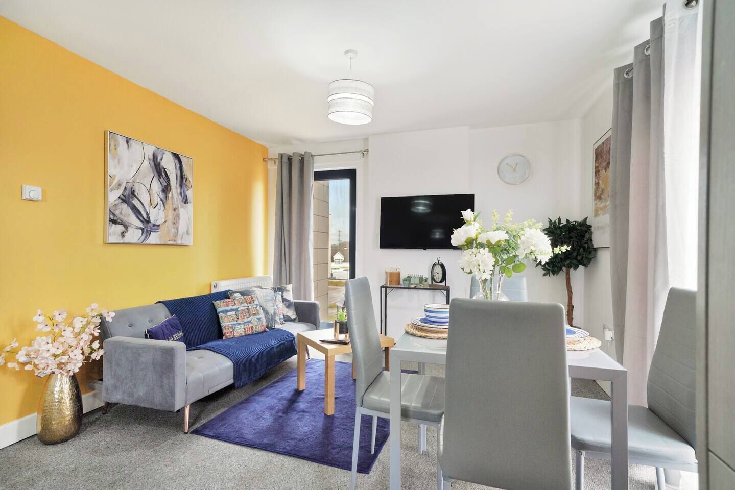 A modern living room with a sofa, dining table, and chairs. There is a flat-screen TV mounted on the wall, a clock, and a vase with flowers.