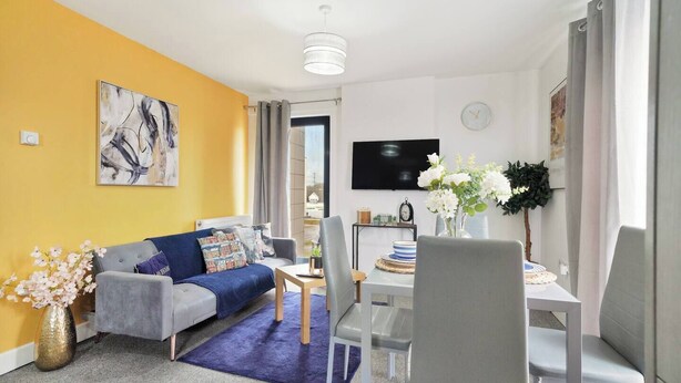 Un salón moderno con sofá, mesa de comedor y sillas. Hay un televisor de pantalla plana en la pared, un reloj y un florero con flores.