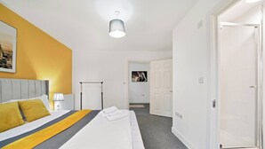 Habitación de hotel con cama, mesita de noche, lámpara, radiador, puerta y pasillo que conduce a un baño.