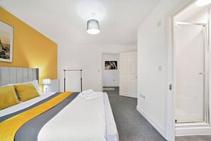 Quarto de hotel com uma cama, mesa de cabeceira, luminária, radiador, porta e corredor que dá para um banheiro.