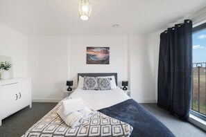 Ein Hotelzimmer mit einem Bett, zwei Nachttischlampen, einem Bild an der Wand, einem Fenster mit Vorhängen und einem weißen Schrank mit einer Pflanze.