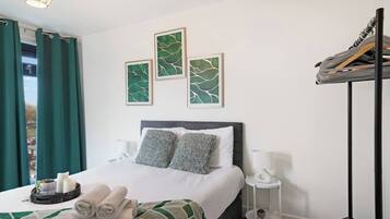 A hotel room with a bed, pillows, a bedside table, a lamp, a wall-mounted shelf, and a window with curtains.