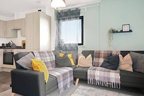 A modern living room with a sofa, a dining area with a kitchen in the background, and a large window with curtains. - Heathrow Gateway Apartments with Private Parking17 (Colnbrook)