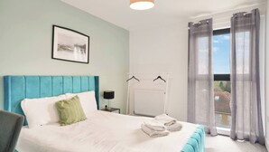 Quarto com cama, mesa de cabeceira, radiador e janela com cortinas.