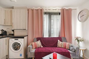 Ein gemütlicher Wohnraum mit Waschmaschine, einer Couch mit roter Decke, einer Uhr an der Wand und einem kleinen Tisch mit Blumen.