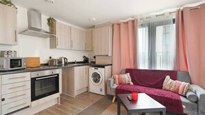 Un ambiente compatto con angolo cottura, lavatrice, divano grigio con coperta rossa e finestra con tende.